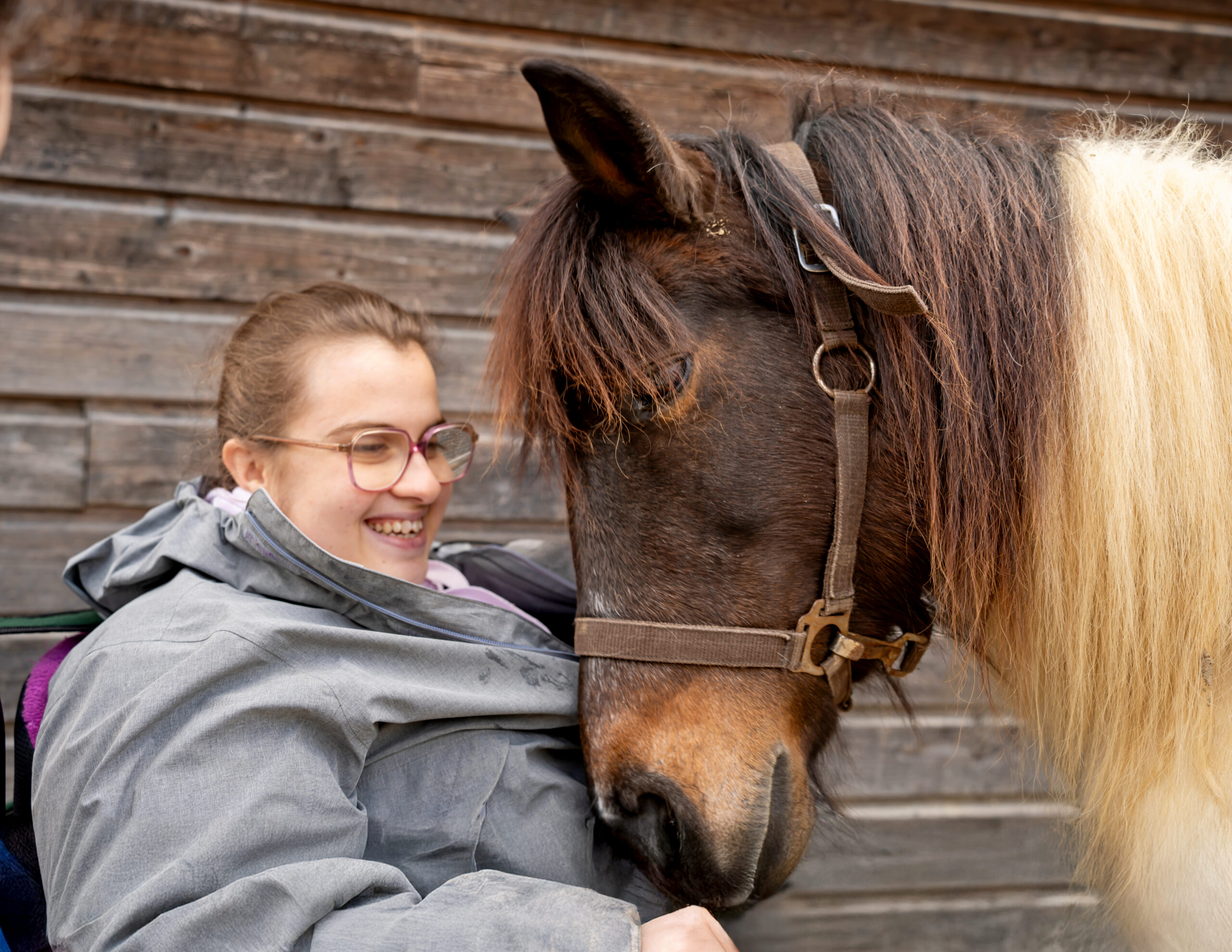 Photo portait d'une jeune fille souriante en fauteuil roulant en train de caresser la tête d'un cheval pendant l'atelier d'équi-handi au mas de rivet à Barjac.