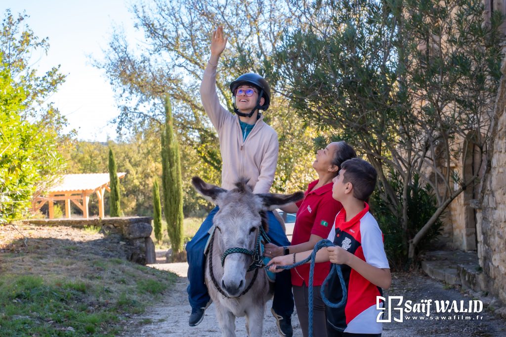 Jeune homme en situation de handicap souriant à dos de mulet accompagnée par une monitrice spécialisée en équi-handi et une autre jeune personne au mas de rivet à Barjac dans le Gard.