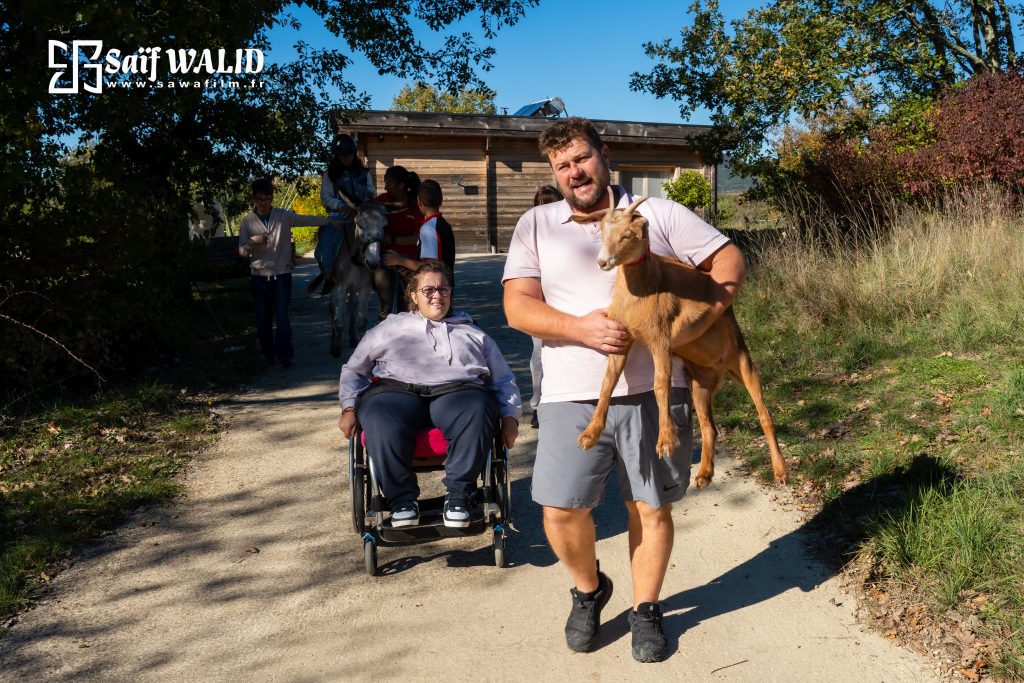Groupe de personnes avec au premier plan moniteur fermette LEX à Barjac portant une chèvre, suivi d'une jeune fille en fauteuil roulant.